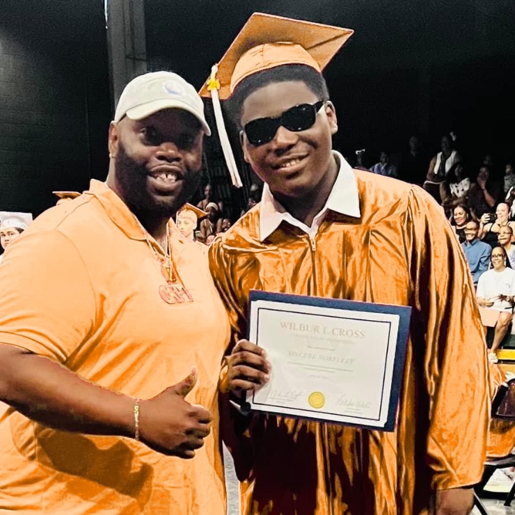 A graduate in cap and gown holding his diploma beside his mentor